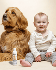 Fluffy brown dog sits next to a baby in light clothing against a neutral backdrop. A Puppy Shampoo bottle stands on the floor in front of them.