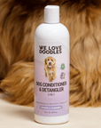 The We Love Doodles conditioner stands upright on a light surface with curly brown dog fur filling the background. The purple label and logo are clear and readable.