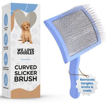 Light blue curved slicker brush with dense stainless steel pins shown beside a We Love Doodles box. A small tag on the handle reads removes tangles, knots and mats.