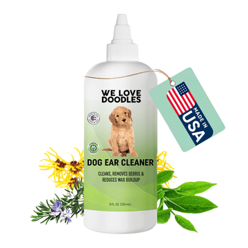 White squeeze bottle labeled We Love Doodles Dog Ear Cleaner on a green label with a puppy image. A Made in USA tag hangs from the cap and sprigs of herbs sit around the bottle on a white background.
