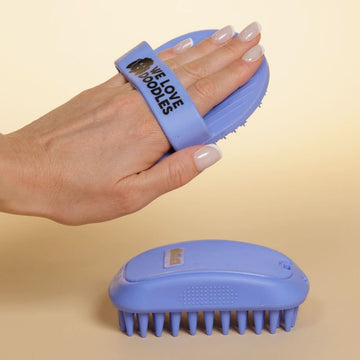A hand holds a blue silicone bath brush with a finger strap. A second blue brush with long tapered pins sits on a beige surface.