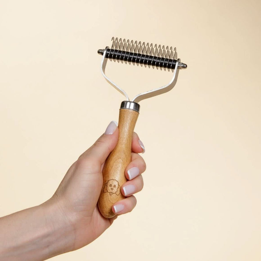 Close view of the dematting comb held in a hand against a light background showing the curved blade rows and wood handle logo.